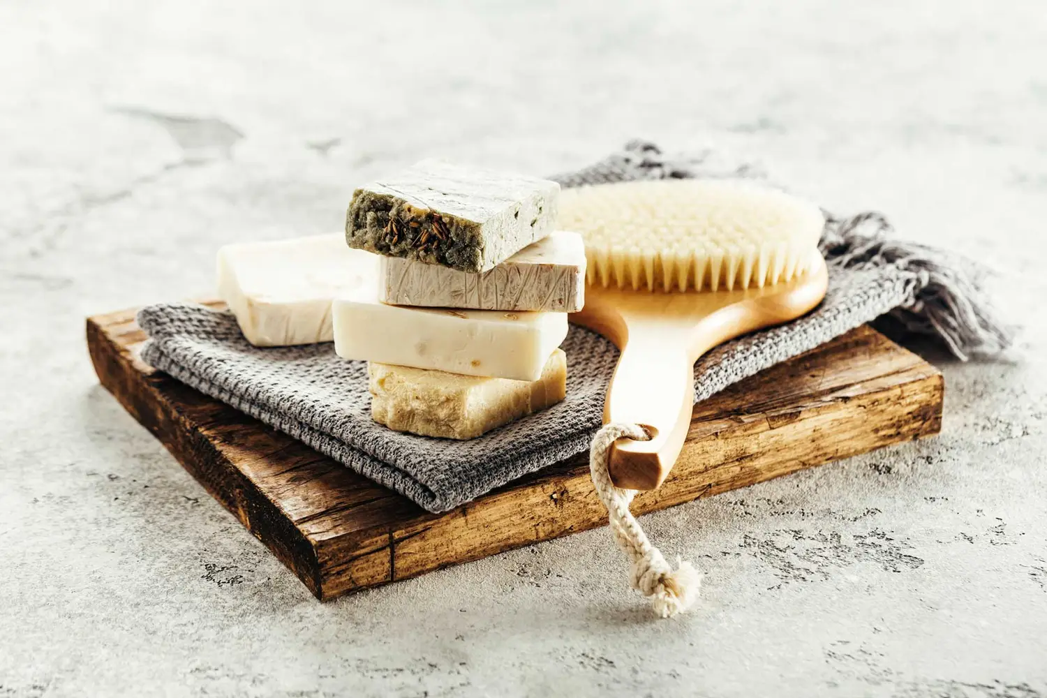 A bar of natural soap with visible herbs and a wooden brush with natural bristles, resting on a rustic wooden table.
